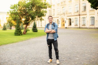 Successful student is standing with backpack behind park near campus. Smile young man standing against university on the background. Education, university, college, studying, course concept