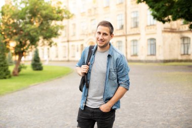 Successful student is standing with backpack near campus on the university background. Smiling young man with bag standing in park. Education, university, college, studying, course concept