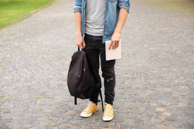 Front view of stylish man stand with book, waiting. Legs student in sneakers, standing on walkway. Man legs in yellow sneakers, wearing grey t-shirt, black skinny jeans at university campus in autumn.