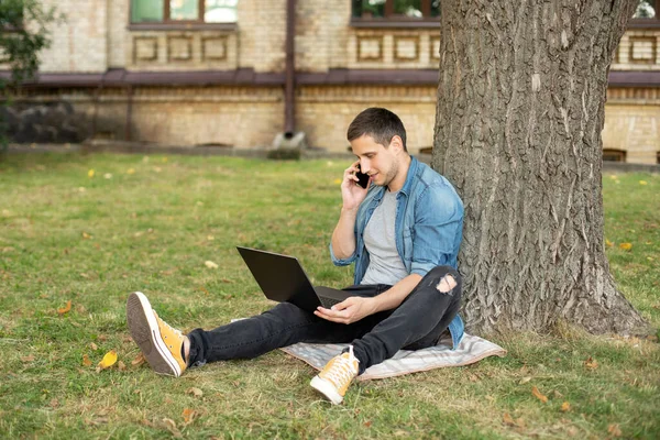 Business man sit on lawn work outdoors on computer and talking on the phone in a park. Young man student with laptop on lesson sit in grass at university campus. education and Remote working concept