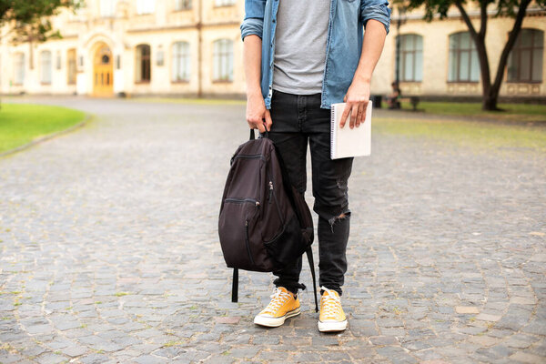 Man legs in yellow sneakers, wearing grey t-shirt, black skinny jeans at university campus in autumn. Front view of stylish man stand with book, waiting. Legs student in sneakers, standing on walkway.