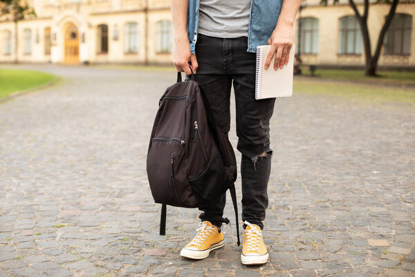 Man legs in yellow sneakers, wearing grey t-shirt, black skinny jeans at university campus in autumn. Front view of stylish man stand with book, waiting. Legs student in sneakers, standing on walkway.