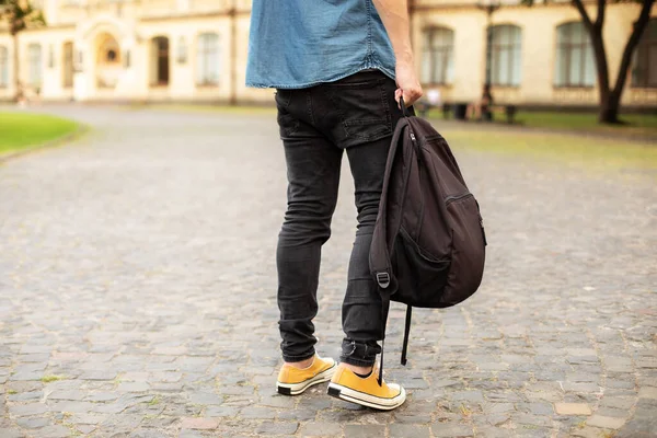 Man legs in yellow sneakers, denim shirt, black jeans at university campus. Back view of stylish man stand with book, waiting. Legs student in sneakers, standing on walkway. education concept