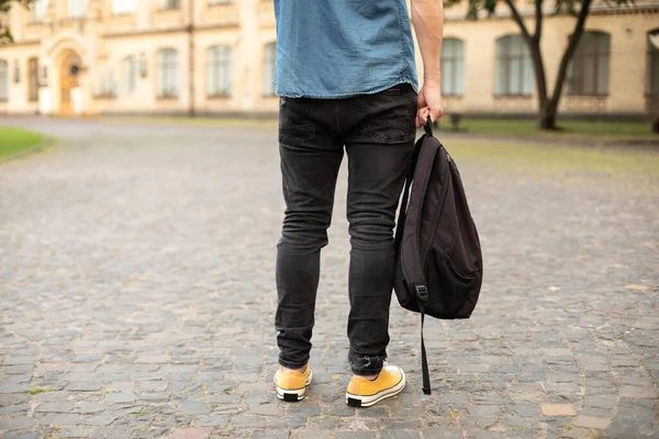 Man legs in yellow sneakers, denim shirt, black jeans at university campus. Back view of stylish man stand with book, waiting. Legs student in sneakers, standing on walkway. education concept