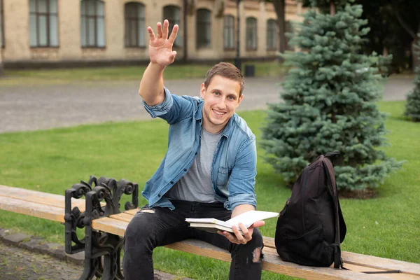 smiling emotional man outdoors looking aside waving hello. Young man with book sit on bench at university campus. Business man work outdoors in a park. education concept. education and Remote working