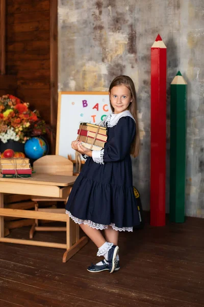 School Girl in dress holds a stack of books. Back to school.  Happy smiling Little girl with books stand near of chalkboard. Kid is studying in the classroom on the background of school board.