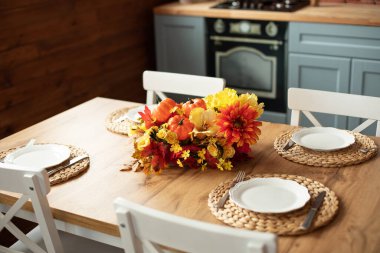 Cozy table setting with fall decor, flowers and utensils in dining room. Bouquet of fall flowers. Cozy kitchen interior. Festive table with wicker napkins, white plates and cutlery on wooden table.