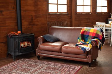 Interior cozy living room with wood burning stove inside, comfortable sofa, rustic wooden walls and carpet on floor. Black modern Cast iron wood stove at home. Fragment of interior of country house. 