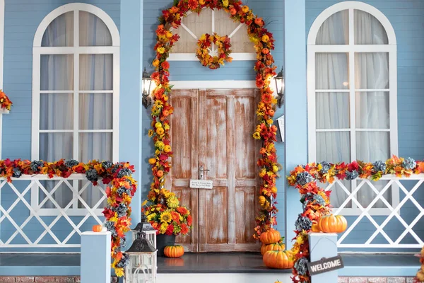 Cozy wooden porch of the house with pumpkins in fall time. Halloween design home with yellow fall leaves and lanterns. House entrance staircase decorated for autumn holidays, fall flowers and pumpkins