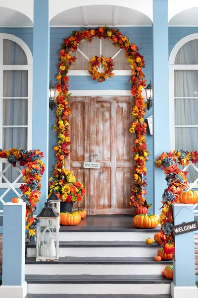 House entrance staircase decorated for autumn holidays, fall flowers and pumpkins. Cozy wooden porch of the house with pumpkins in fall time. Halloween design home with yellow fall leaves and lanterns