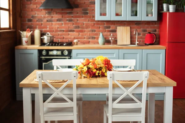 Cozy cuisine decorated with fall decor and table setting flowers and pumpkins. Interior design scandinavian kitchen with utensils, dishes, plates. Stylish dining room with wooden table and chairs.