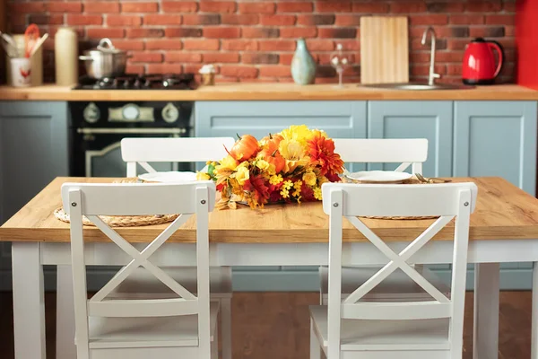 cuisine decorated with fall decor and table setting flowers and pumpkins. Scandinavian kitchen with utensils, dishes, plates. Stylish dining room with wooden table and chairs.