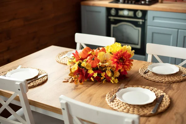 Cozy table setting with fall decor, flowers and utensils in dining room. Bouquet of fall flowers. Cozy kitchen interior. Festive table with wicker napkins, white plates and cutlery on wooden table.