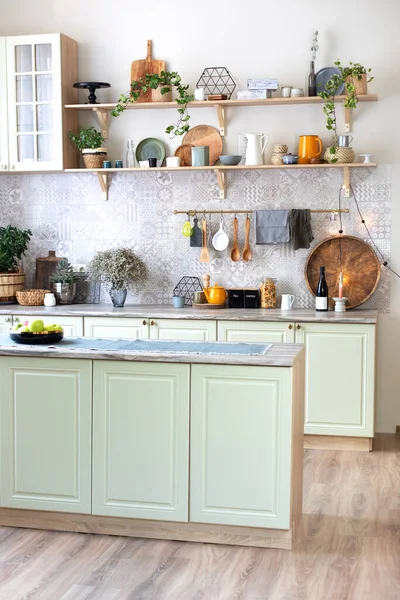 Green wooden kitchen interior with wooden shelf and cozy decoration. Stylish cuisine with flowers in vase. Modern home decor. Kitchen utensils, dishes and plates. Kitchen island in dining room.