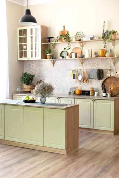 Modern home decor. Green wooden kitchen interior with wooden shelf and cozy decoration. Stylish cuisine with flowers in vase. Kitchen utensils, dishes and plates. Kitchen island in dining room.