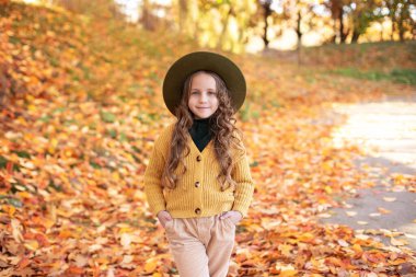 Sonbahar arka planında şapkalı ve sonbahar elbiseli mutlu küçük kız. Sonbahar parkında gülümseyen bir çocuk oynuyor. Küçük kız altın yapraklarla oynarken eğleniyor. Güzel bir kızın sonbahar portresi.