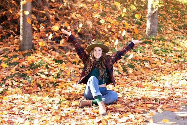Young girl in hat and autumn clothes playing with golden fall leaves in hand. Happy girl in green hat and plaid jacket on an autumn background. A smiling girl walking in fall park.