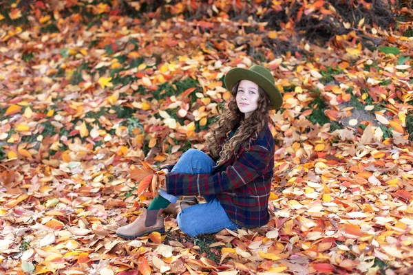 Young girl in hat and autumn clothes with golden fall leaves in hand. Happy little girl in green hat and plaid jacket on an autumn background. A smiling girl walking in fall park.