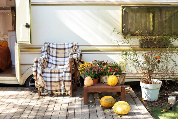 Wooden armchair near outside caravan trailer decorated for Halloween. Wooden RV house porch with garden furniture. Campsite in garden. Interior cozy yard campsite with fall flowers potted and pumpkins