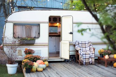 Sonbahar çiçekleri saksısı ve balkabaklarıyla iç taraftaki kamp alanında. Karavan karavanının yanındaki tahta koltuk Cadılar Bayramı için dekore edilmiş. Bahçe mobilyalarıyla ahşap karavan evi verandası. Bahçede kamp yeri