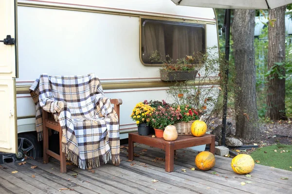 Campsite in garden. Interior cozy yard campsite with fall flowers potted and pumpkins. Wooden armchair near outside caravan trailer decorated for Halloween. Wooden RV house porch with garden furniture