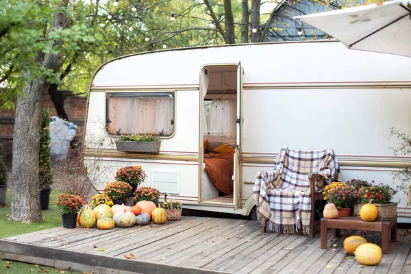 Interior cozy yard campsite with fall flowers potted and pumpkins. Wooden armchair near outside caravan trailer decorated for Halloween. Wooden RV house porch with garden furniture. Campsite in garden