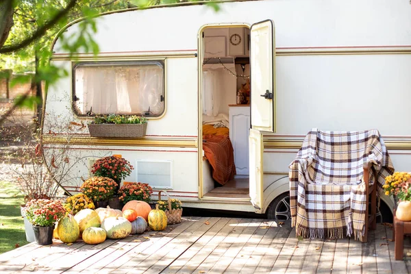 Halloween design home. Interior cozy yard campsite with fall flowers potted and pumpkins. Wooden RV house porch with garden furniture. Campsite in garden. Wooden armchair near outside caravan trailer