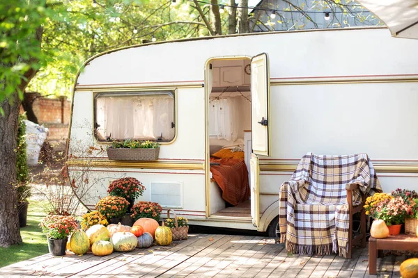 Wooden RV house porch with garden furniture. Campsite in garden. Interior cozy yard campsite with fall flowers potted and pumpkins. Wooden armchair near outside caravan trailer decorated for Halloween