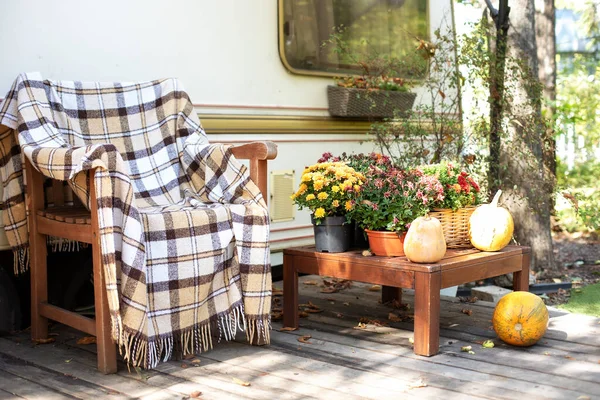 Wooden armchair near outside caravan trailer decorated for Halloween. Wooden RV house porch with garden furniture. Campsite in garden. Interior cozy yard campsite with fall flowers potted and pumpkins