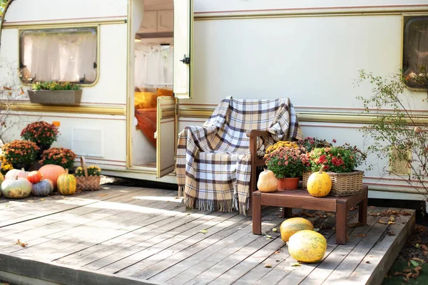 Wooden armchair near outside caravan trailer decorated for Halloween. Wooden RV house porch with garden furniture. Campsite in garden. Interior cozy yard campsite with fall flowers potted and pumpkins