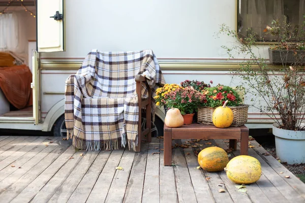 Wooden armchair near outside caravan trailer decorated for Halloween. Wooden RV house porch with garden furniture. Campsite in garden. Interior cozy yard campsite with fall flowers potted and pumpkins