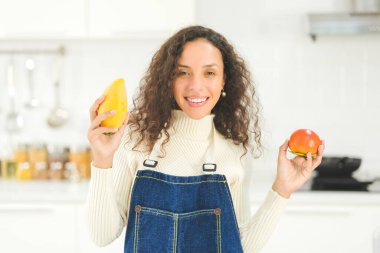 Güzel bir kadının portresi gülümsüyor ve parlak beyaz mutfakta mango ve domates tutuyor. Eş ya da ev hanımı aile için sağlıklı yemek pişirmeye hazırlanıyor. Yemek siparişi..