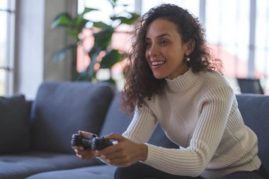 Young African American woman is having fun playing video games with a single joystick on sofa at home. Social distancing during the COVID-19 epidemic.