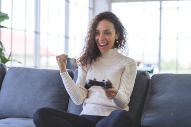 Young African American woman is having fun playing video games with a single joystick on sofa at home. Social distancing during the COVID-19 epidemic.