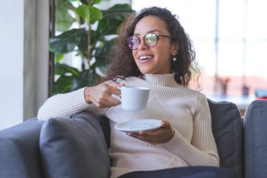 Portrait of beautiful young African American woman wearing eye glasses and relaxing and drinking coffee on sofa in holiday at home. business women and freelancers working from home to spread coronavirus covid-19