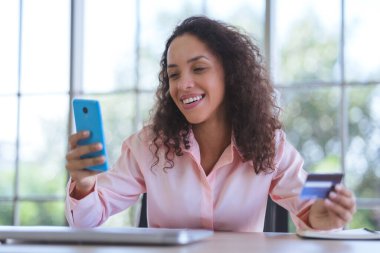Genç Latin kadın gülümsüyor ve online alışveriş için akıllı telefon kullanıyor. İş kadını ve evden bağımsız çalışıyor ve elektronik ödeme için kredi kartı tutuyor. Çevrimiçi mağazada mutlu alışveriş hizmetleri.