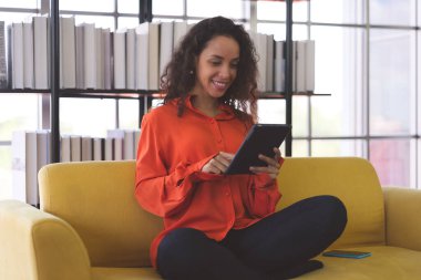 Portrait of a young black woman wearing orange shirt smiling and using tablet for shopping online, leaning online, and video call on sofa at home. Business Woman work from home in spreading of coronavirus covid-19.