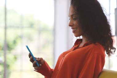 Turuncu tişört giyen, gülümseyen, akıllı telefon kullanan, online alışveriş yapan ve evdeki kanepede iletişim kuran güzel siyahi bir kadın. İş kadını ve serbest çalışan Coronavirus covid-19 'un yayılması için evden çalışıyor..