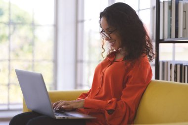 Portrait of a young black woman wearing orange shirt smiling and using laptop for shopping online, leaning online, and video call on sofa at home. Business Woman work from home in spreading of coronavirus covid-19.