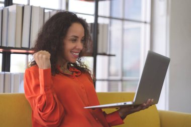Successful young black woman wearing orange shirt exciting and using laptop for video call on sofa at home. Business Woman and freelance work from home in spreading of coronavirus covid-19.