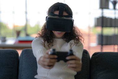Young African American woman wearing virtual reality glasses is playing vr video games with joystick on sofa at home. Stay at home and self-quarantine in spread of coronavirus covid-19