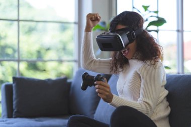 Young African American woman wearing virtual reality glasses is playing vr video games with joystick on sofa at home. Stay at home and self-quarantine in spread of coronavirus covid-19