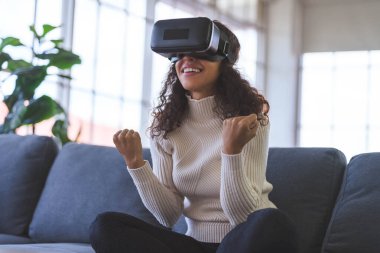 Young African American woman wearing virtual reality glasses is playing vr video games on sofa at home. Stay at home and self-quarantine in spread of coronavirus covid-19