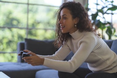 Young African American woman is having fun playing video games with a single joystick on sofa at home. Social distancing during the COVID-19 epidemic.
