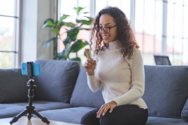 Young African American women wearing eyeglasses live streaming training on the sofa through the application from smartphone to social media during the coronavirus outbreak.