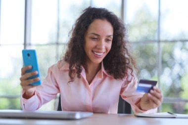 Genç Latin kadın gülümsüyor ve online alışveriş için akıllı telefon kullanıyor. İş kadını ve evden bağımsız çalışıyor ve elektronik ödeme için kredi kartı tutuyor. Çevrimiçi mağazada mutlu alışveriş hizmetleri.