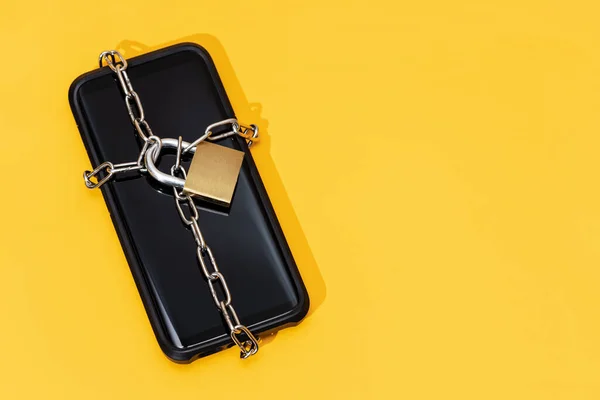 Phone security concept. Smartphone locked with chain and padlock on yellow background. Mobile security and data privacy. Copy space