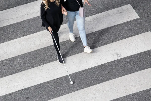 Scene of a Blind woman walking on zebra crossing helped by another person using her white cane. Help in the early stages of blindness