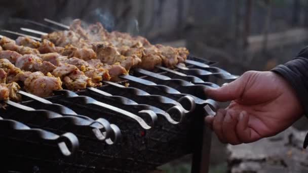 Egy férfi nyárson süt barbecue-t faszén grillen az utcán. Grillező a szabadban. Piknik a természetben. Pörgetés nyársak grillezett hús közelkép — Stock videók