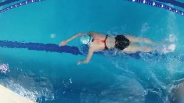 Eşofmanlı, dalış gözlüklü, mayo takan kadın yüzücü havuzda antrenman yapıyor. Sporcu serbest bir şekilde yüzer. Geniş açı, 4k..
